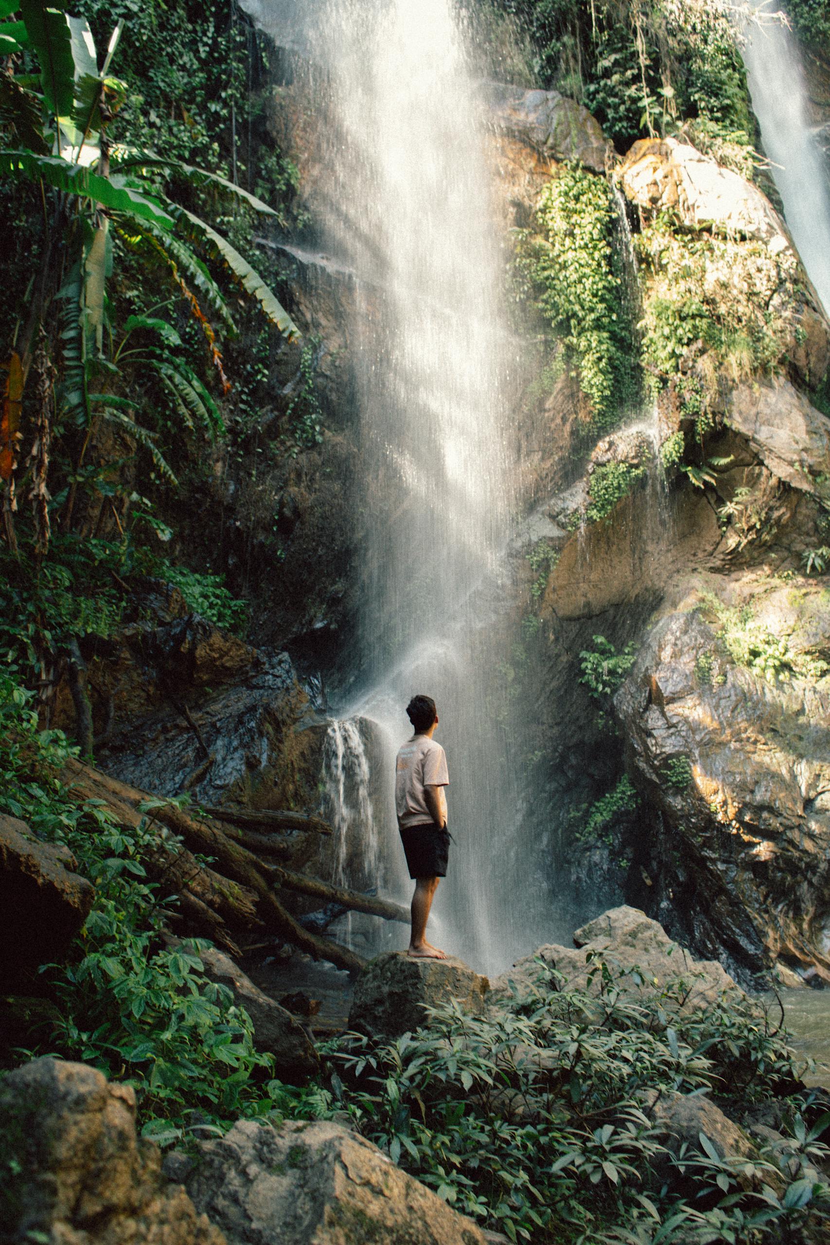A man stands in awe at a lush waterfall surrounded by greenery in Chiang Mai, Thailand.